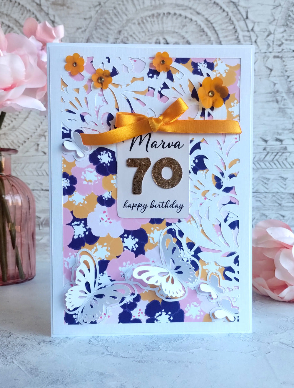Birthday card with floral design, yellow ribbon, and '70 happy birthday' tag on a light background.