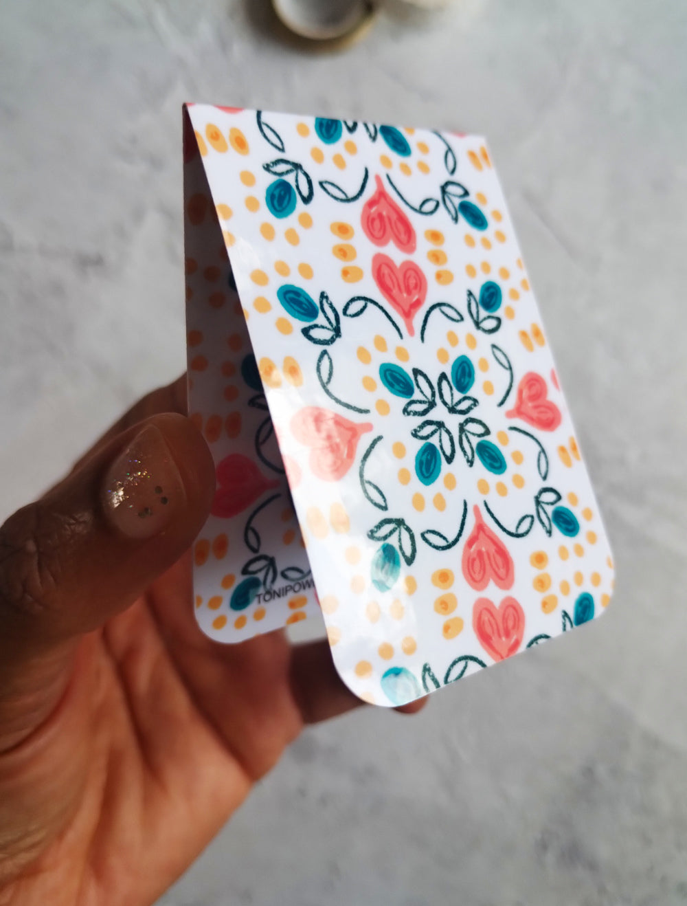 Hand holding a colourful, glossy Summa Love magnetic bookmark with heart pattern design